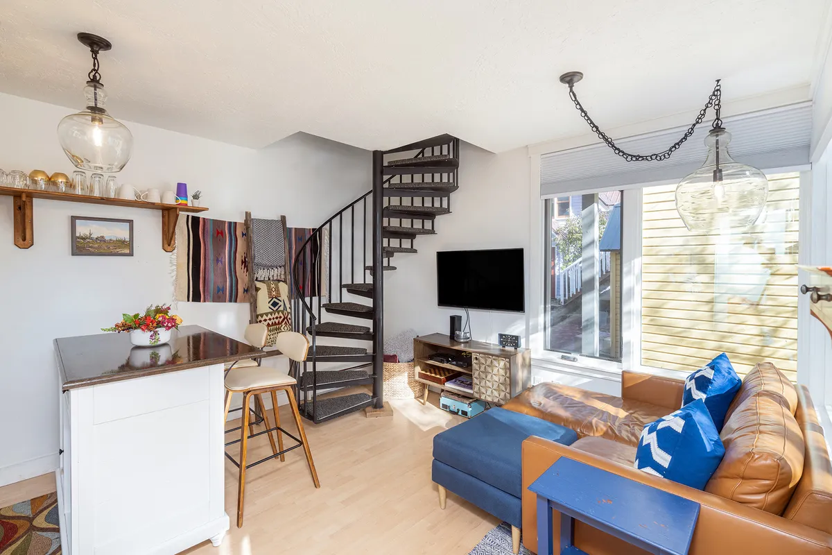 The image shows a cozy, inviting living space featuring a modern spiral staircase, a plush sofa perfect for relaxing, a sleek kitchen island ideal for intimate meals or gatherings, and a flat-screen television for entertainment. Natural light fills the room, creating a warm and welcoming atmosphere.
