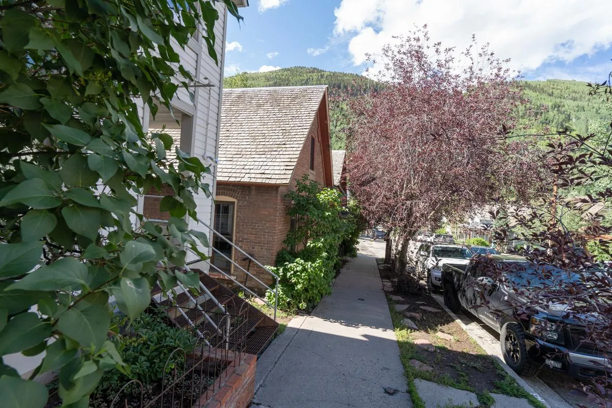 A picturesque exterior street scene featuring lush green foliage, a brick wall, colorful trees, a sidewalk, and distant mountains. The welcoming atmosphere invites leisurely strolls and social connections in the heart of the community.