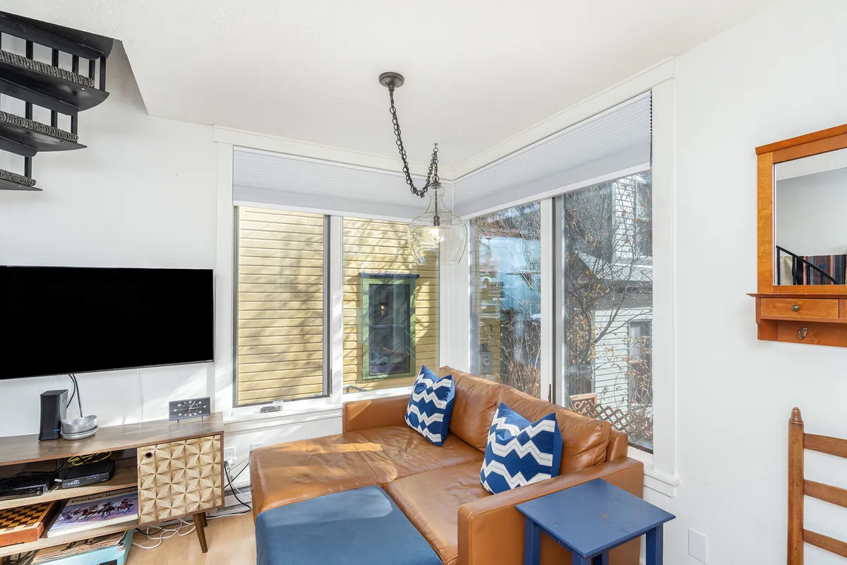 This image showcases a comfortable living area featuring a leather sofa with decorative pillows, a wall-mounted TV, and a wooden shelf with a mirror. The room is bright, inviting, and well-suited for relaxing, socializing, or enjoying a quiet afternoon.
