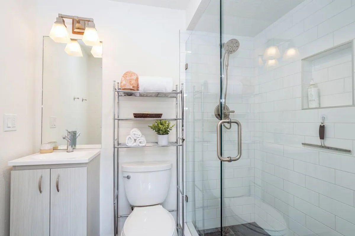 A modern bathroom with a glass shower enclosure, mirrors with built-in lights, and a shelf with towels and a plant. The serene and refreshing decor hints at comfort and luxury.