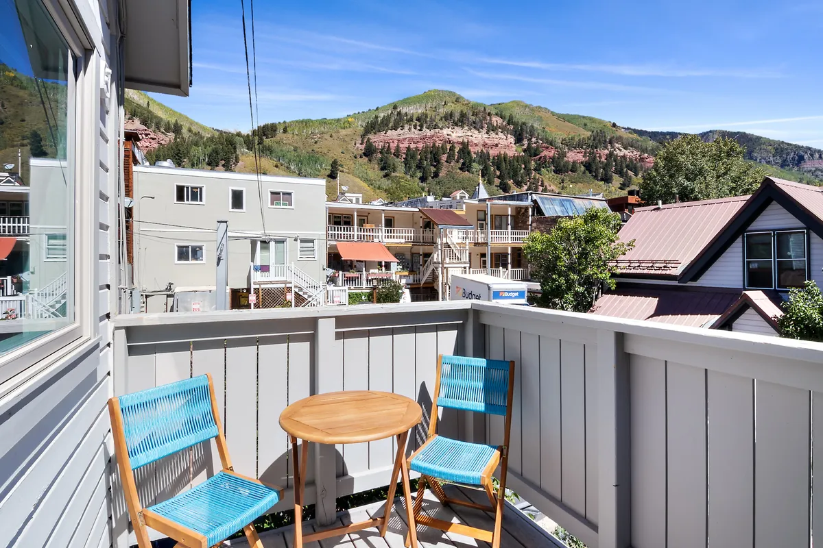 This image displays the balcony seating area with stunning mountain views beyond, showing a serene spot ideal for morning coffee or relaxation. The surrounding buildings hint at the sense of place.