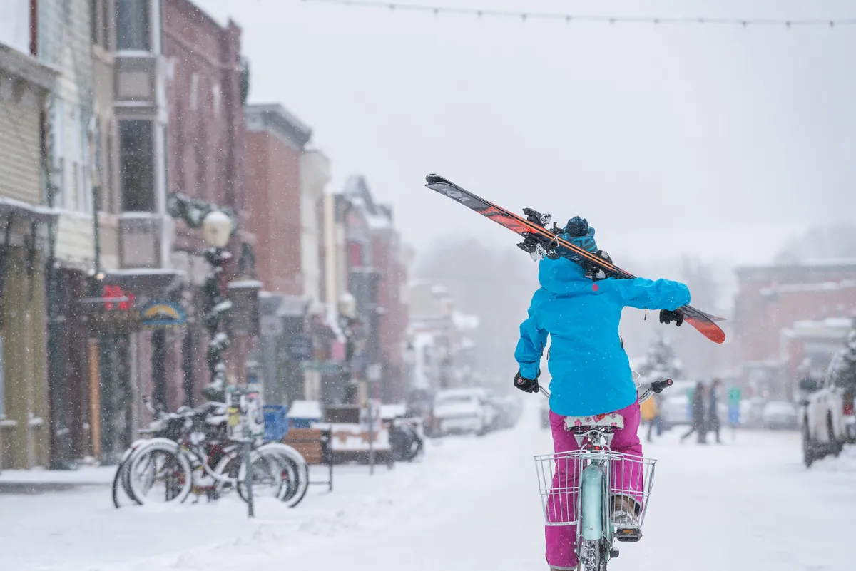 A wintery street scene with a bicyclist carrying skis, snow-covered sidewalks, and colorful storefronts. The photo blends elements of biking and skiing, two core activities referenced in the section.