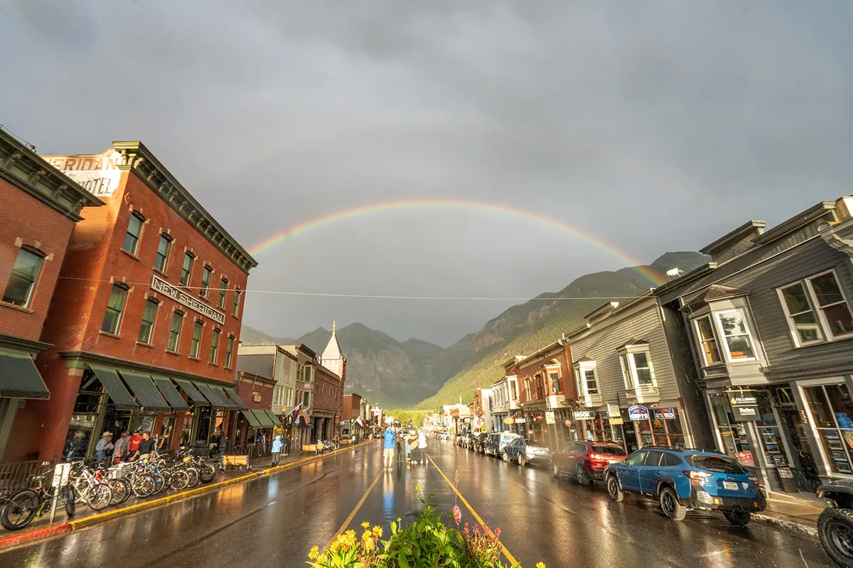 A view down a historic street lined with beautiful old buildings, with a rainbow arching across a wet street and bicycles parked along the sidewalk. Mountains rise in the background, and the atmosphere feels lively and uplifting—perfect for an event-filled day in downtown Telluride.