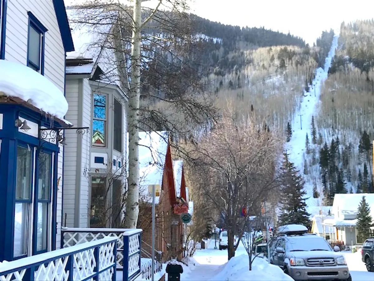 A snowy street lined with colorful houses, a visible ski lift trail in the background, and a mountain backdrop. The inviting scene shows people walking and the vibrancy of the local neighborhood.