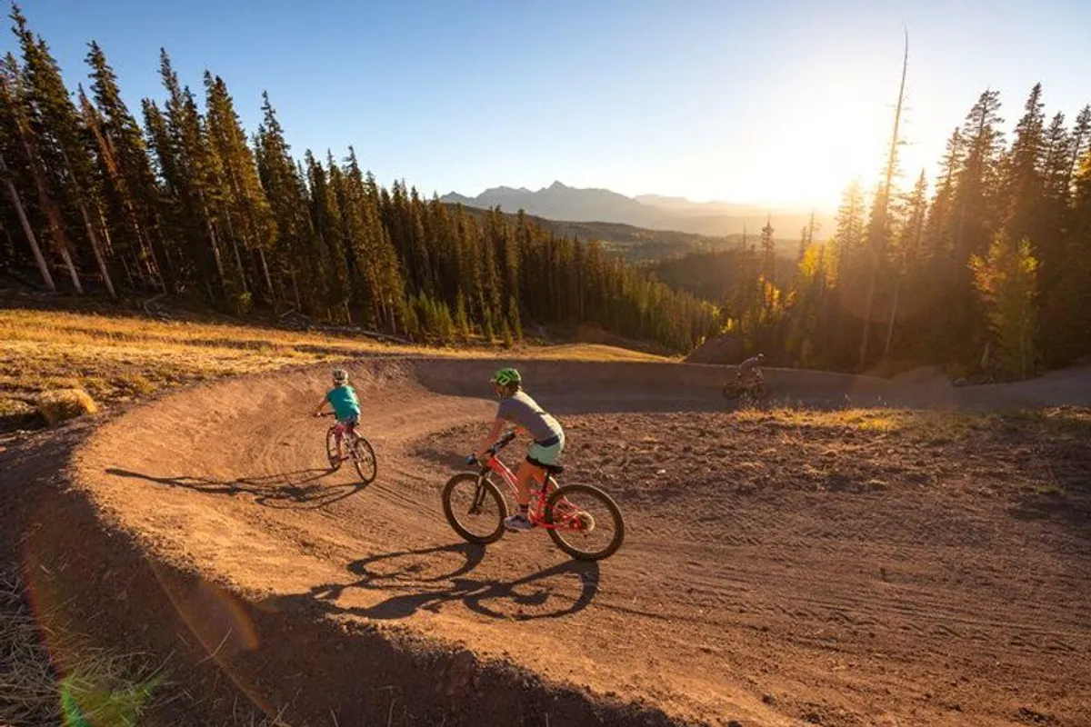 An exterior scene with mountain bikers riding along a curved dirt trail surrounded by lush pine trees and a stunning mountain backdrop. The image conveys action, movement, and outdoor excitement.