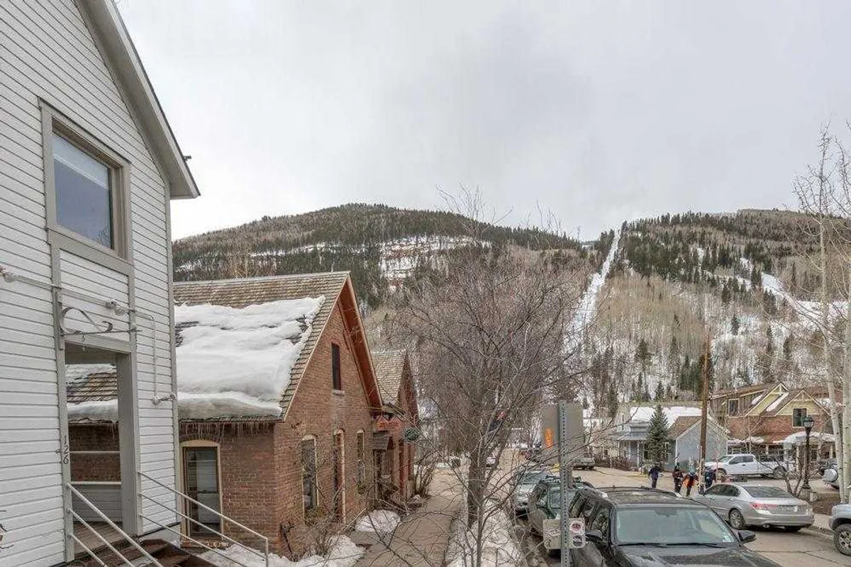 This exterior image captures Telluride's historic brick buildings lining a tree-lined street, with fresh snow on the ground and the majestic mountains rising in the background. The festive atmosphere and visible storefronts hint at an active local scene, perfect for strolling, shopping, and discovering local cuisine.