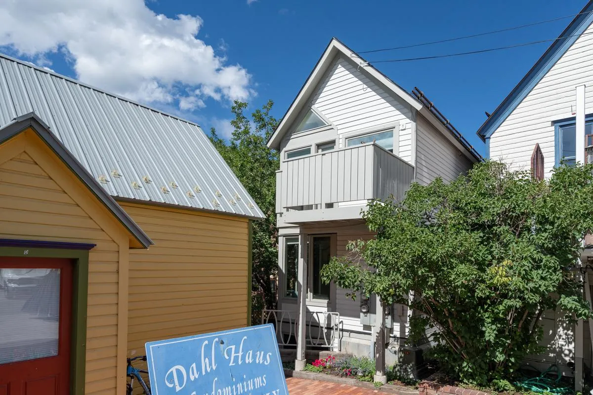 This exterior shot features two distinct buildings, one yellow and one white, with the sign 'Dahl Haus Condominiums' and lush green foliage framing the scene. The architecture hints at a vibrant, welcoming neighborhood ideal for festival visitors who want to be in the midst of the action.