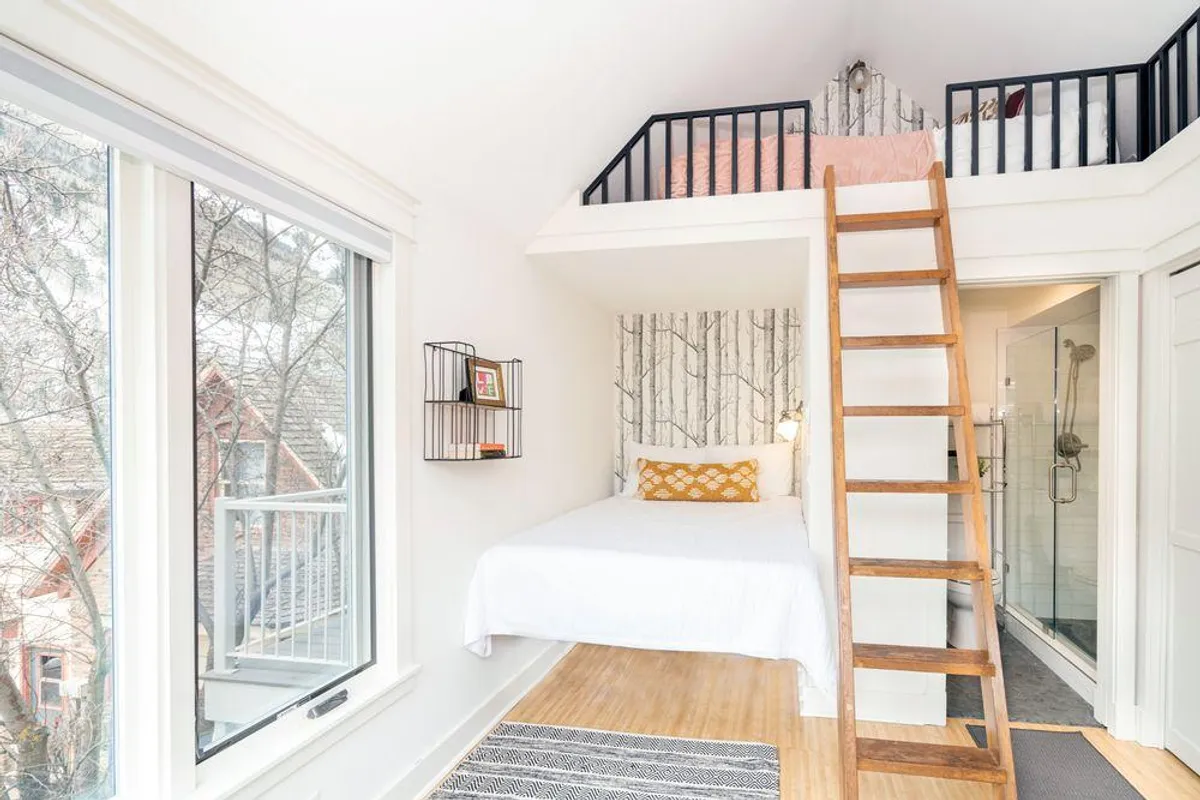 The image features the open loft with a queen bed, whimsical aspen tree wallpaper, a wooden ladder for access, and thoughtful decorative touches. Sunlight filters through the windows, enhancing the cozy and intimate feel.