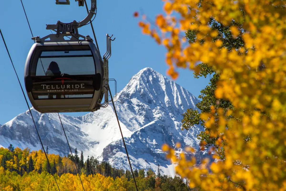 The image features a gondola mid-ride with a snow-capped mountain in the background and brilliant autumn trees below. The scene captures the transition between seasons and highlights the close proximity of the gondola to the mountainous terrain.