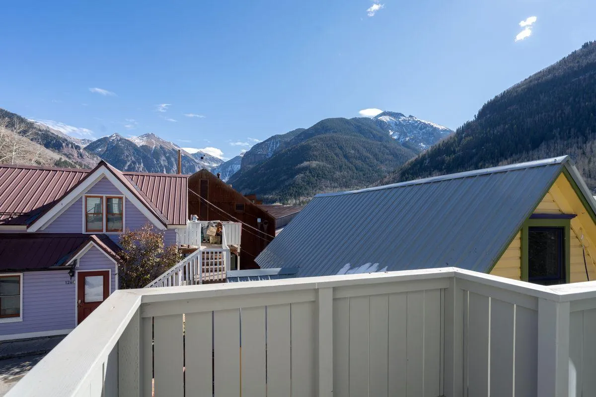 A panoramic view featuring a mountain backdrop framed by colorful rooftops beneath a clear blue sky. The perspective is likely from a balcony or an elevated position, inviting viewers to imagine relaxing and soaking in Telluride's natural beauty.