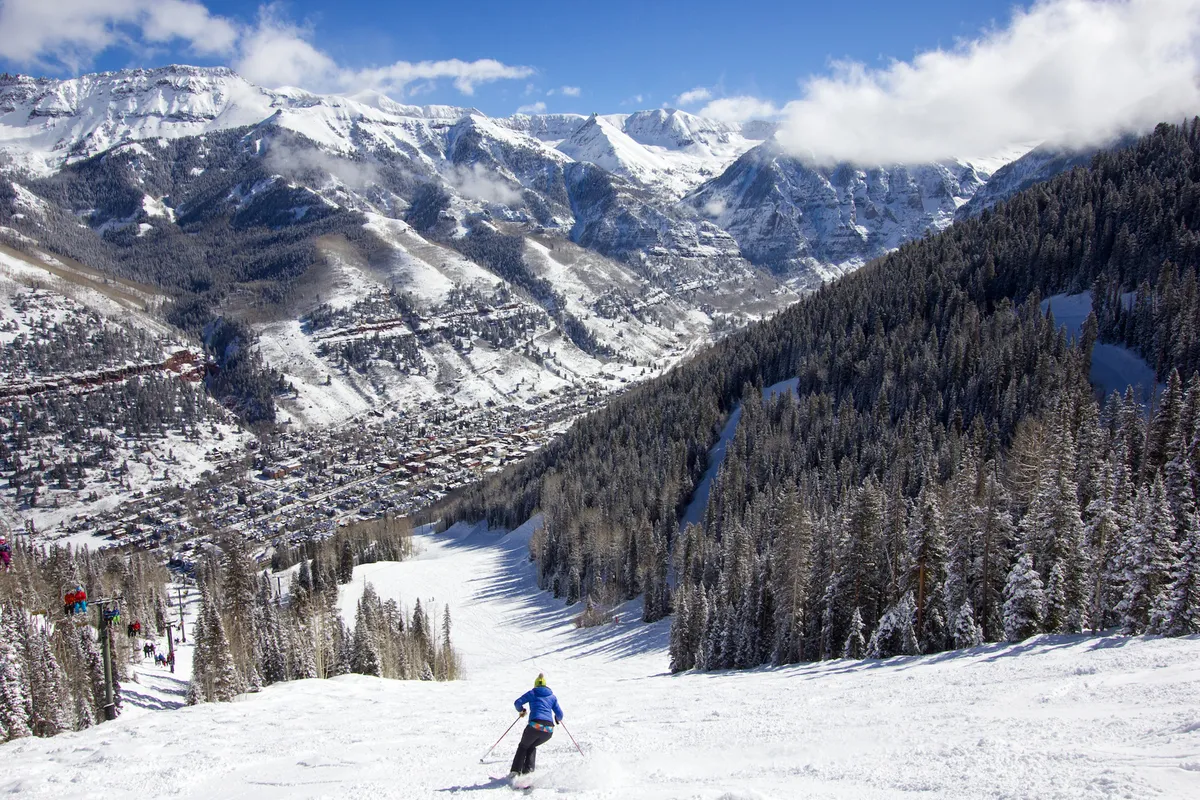 A skier descends a snow-covered slope overlooking a picturesque mountain town nestled in a valley with pine trees and mountains surrounding. The wide view encompasses both the thrill of the slopes and the proximity to the town below.